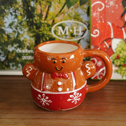 A smiling gingerbread man shaped mug. It is brown,  with a red bow, whites buttons, red & white suspenders, and red base with white snowflakes on it. The mug has a brown handle. The mug is on a brown table, with blurred photos and christmas decor in the background.