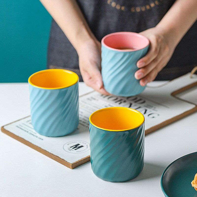 Person's hands holding a blue & pink Sweet Swirl mug with coffee in it. There is a blue & yellow Sweet swirl mug and a green & yellow Sweet Swirl mug next to it.