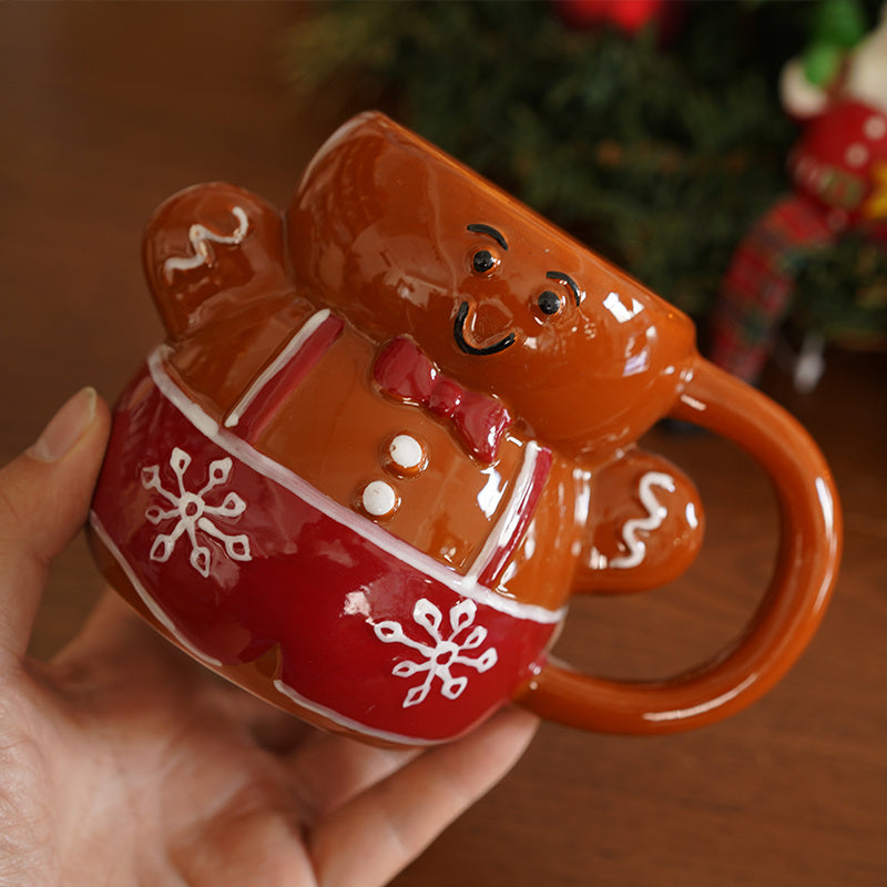 A person's hand holding a A smiling gingerbread man shaped mug. It is brown,  with a red bow, whites buttons, red & white suspenders, and red base with white snowflakes on it. The mug has a brown handle. In the background is a hardwood floor, a and a blurred decorated Christmas tree.