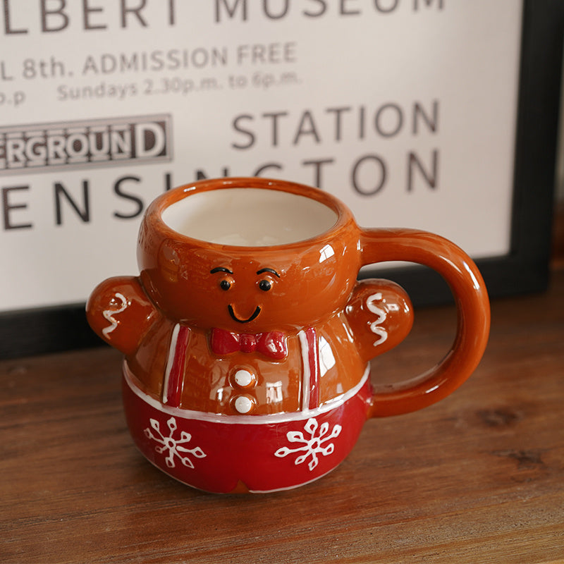 A smiling gingerbread man shaped mug, which is on a brown wooden table. It is brown,  with a red bow, whites buttons, red & white suspenders, and red base with white snowflakes on it. The mug has a brown handle. There is a black and white framed text based poster in the background.