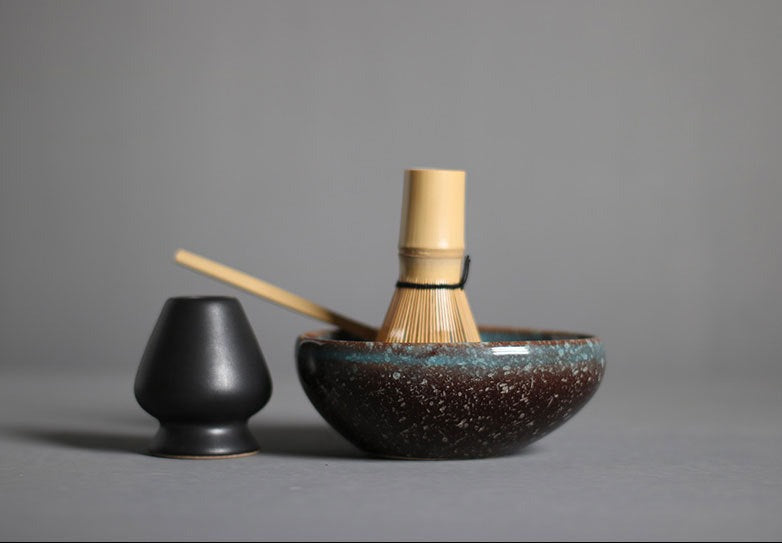 4 Piece Matcha Tea Set With Brown & Blue Speckled Bowl. The matcha whisk and scoop are inside of the bowl and on the left is a black matcha whisk holder.