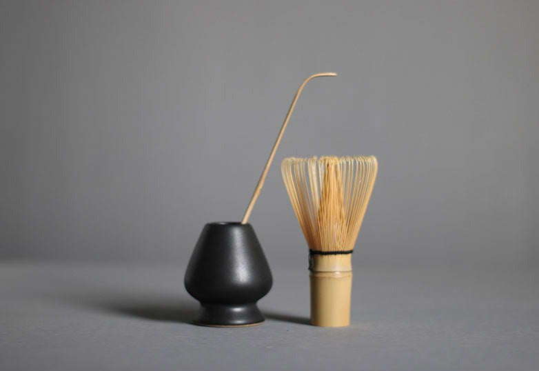 On the left there is a matcha tea whisk holder with a matcha gtea scoop in side of it. On the right there is a matcha tea whisker standing upright, handle side facing down.