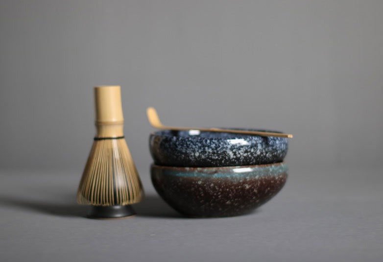 A matcha tea whisk sitting on top on the matcha whisk holder. On the right of them is 2 stacked matcha tea bowls. One in blue & brown speckled (on the bottom) and the other in blue and black speckled (on the top). There is a matcha tea scoop resting on top of the bowls.