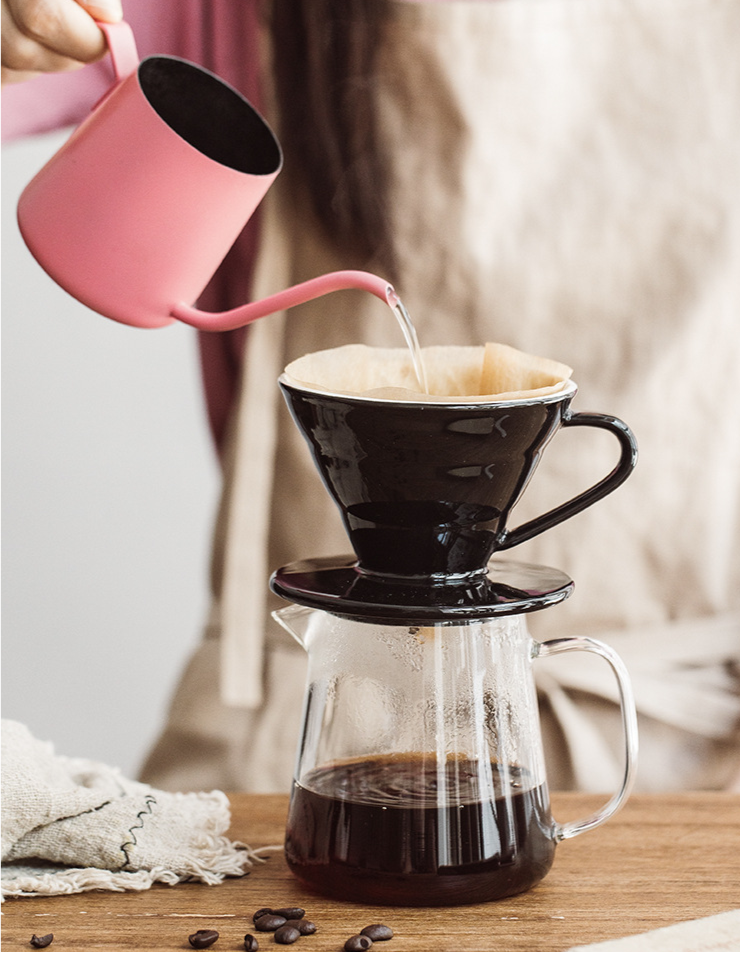 Person pouring hot water from a pink kettle into a black Ceramic Pour Over Coffee Filter which is sitting on top of a clear coffee pot, which is on top of a wooden table. There is a tea towel and coffee beans scattered on the table. There is a beige coffee filter in the ceramic pour over and black coffee in the coffee pot.