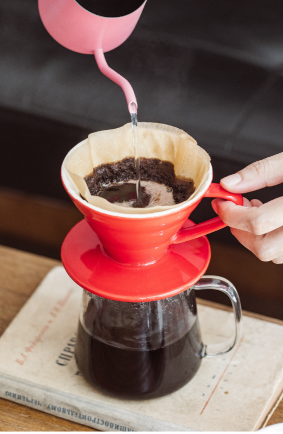 Person pouring hot water from a pink kettle into a red Ceramic Pour Over Coffee Filter which is sitting on top of a clear coffee pot, which is on top of a wooden table.  The coffee pot is sitting on top of a book.. There is a beige coffee filter in the ceramic pour over and black coffee in the coffee pot.