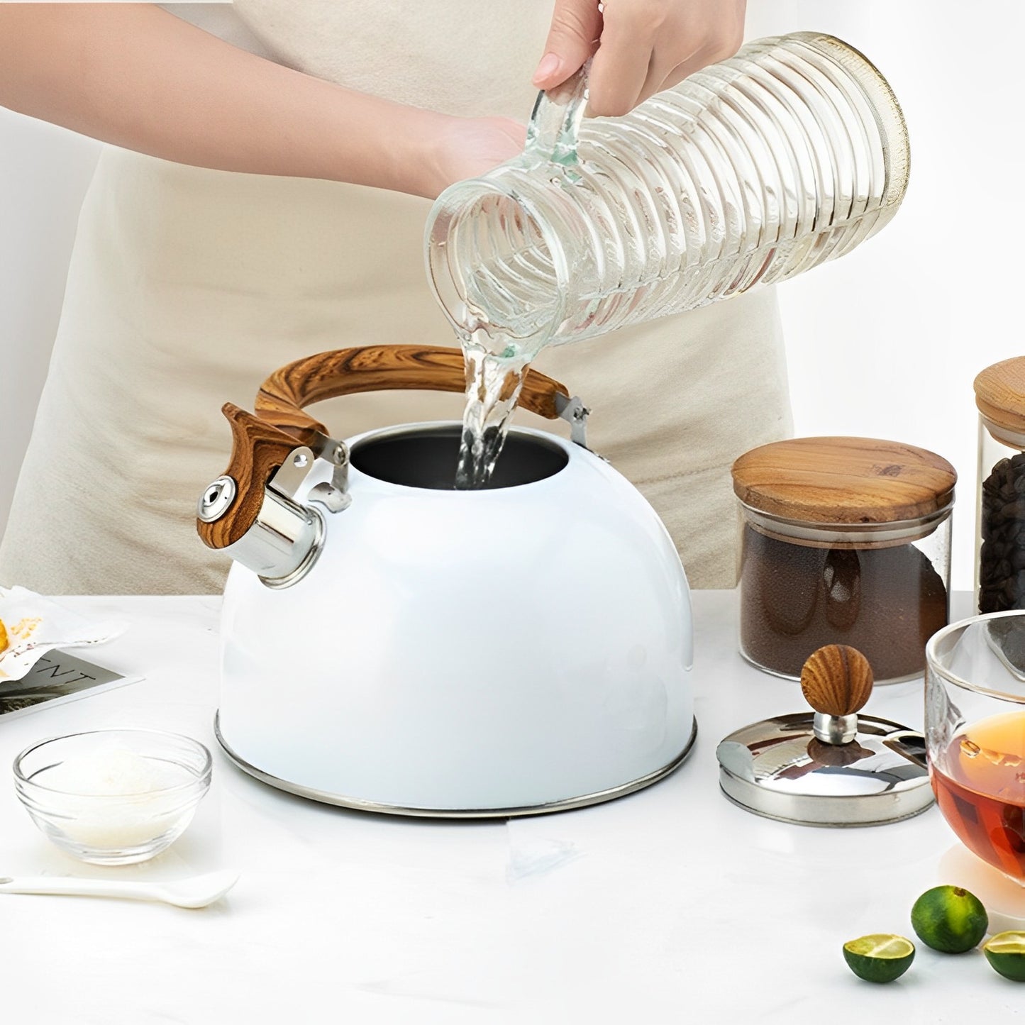 A person pouring water from a glass pitcher/jug into a White Kitchen Kettle with Wooden Handle, that is on top of a tabele next to some jars of coffee & tea, limes, and a clear glass rammican & spoon, and a clear glass mug of black tea.