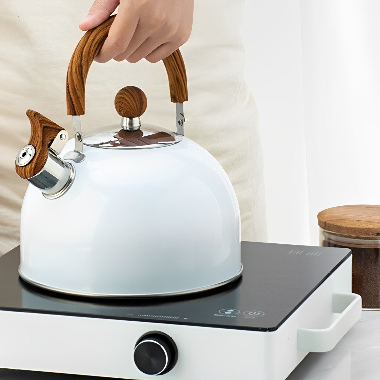 A person holding a White Kitchen Kettle with Wooden Handle which is on top of a portable1 burner electric stove, a top a table with ground coffee in a jug on the table as well.