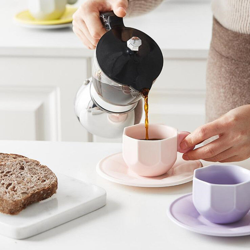 Person pouring coffee from a moka pot into an Octagonal Shaped pink Mug with Macaron Handle on top of a saucer. There is another mug set beside it that is purple, on the bottom right. On the left there is a piece of whole wheat bread on a cutting board. In the background there is another off the same type of mug sitting atop of two stack saucers on a kitchen counter/bench. 