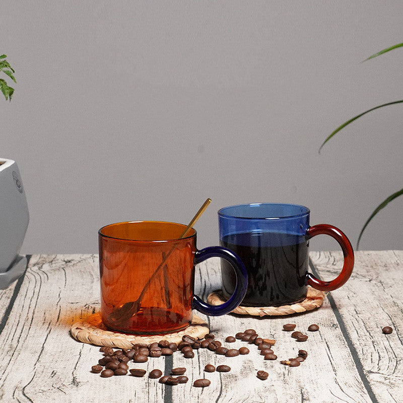 Retro 70s Style Vintage Blue and Amber Colored Glass Mugs on rattan coasters, on a wooden table with coffee beans strewn on the table. Plant leaves on the right and left hand corners of the photo.