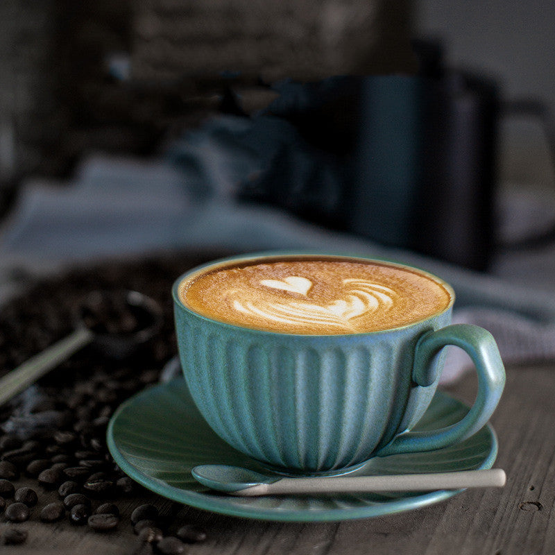 Vintage style ceramic mug and saucer in muted teal blue with coffee coffees and black milk frother in the background, on table. There is a spoon on the saucer as well.