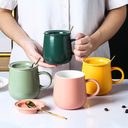 Person holding dark green "Keep 'Er Warm" Mug with Lid and Spoon. There are 3 other mugs with lids in front of them: A light green mud with lid and spoon on the left, a pink mug with the lid off and spoon on the table, and on the right there a yellow mug with lid on and the spoon inside. The are a few coffee beans strewn on the table and a plate on the left hand side of the table.