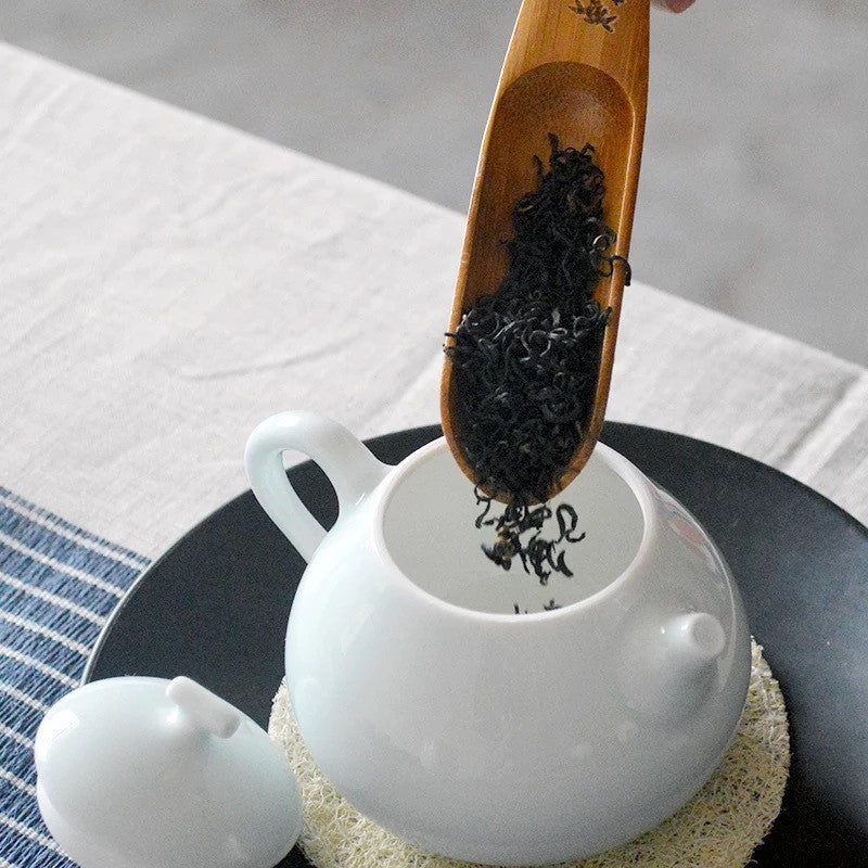 Bamboo Tea Spoon added loose tea leaves to a white kettle on a plate, on top of a table.