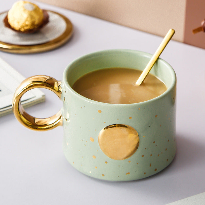 Golden Accented ceramic Mug with Sun Design on table with spoon and chocolate on a plate in the background.