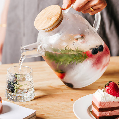 A person's hand pouring fruit and herb infused water from the Clear Glass Pitcher With Bamboo Lid into a glass.