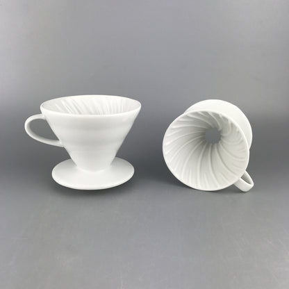 2 white Ceramic Pour Over Coffee Filters on a gray backdrop. The ceramic coffee filter on the left is upright and the ceramic coffee filter on the right is on its side with the top side facing forward so the swirl pattern on the inside of the coffee filter is displayed.