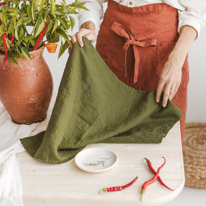 Woman wearing apron, holding Olive Green Tea Towel, with plant, plate with herbs, and chile peppers in the background.