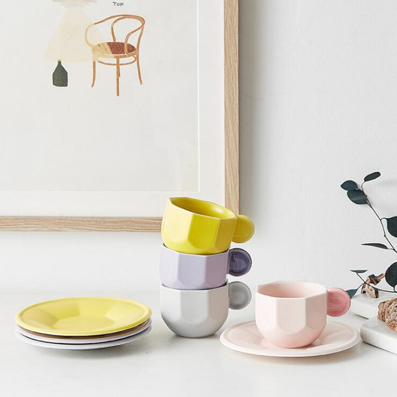 Octagonal Shaped Mug Set with Macaron Handle. Mugs stacked atop each other, plates stacked atop each other, and a mug on top of saucer, and a plant beside it, all on a table. There is a painting of a chair in the background.