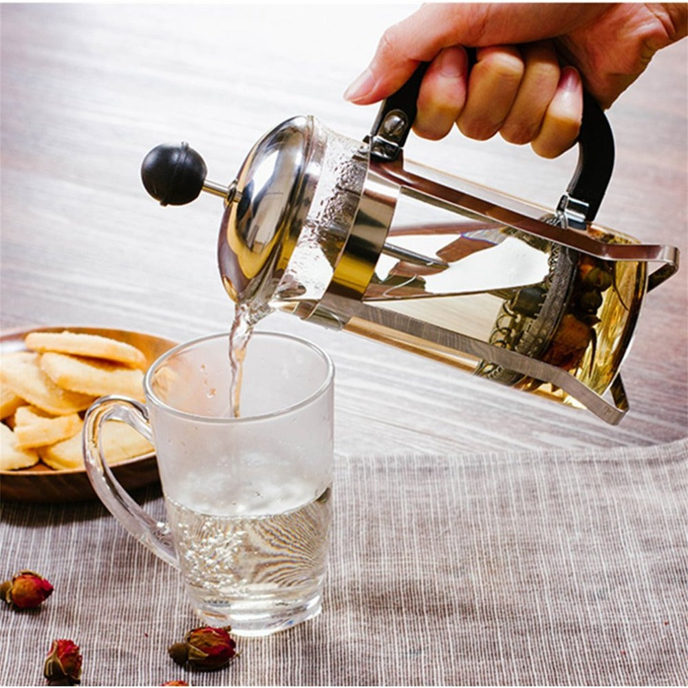 A hand using a MiniBrew Stainless Steel French Press to pour tea into a glass mug, which is on a table (with a beige linen tablecloth) with a bowl of biscuits behind the glass mug (on the left). There are dried rose buds on the front left, surrounding the glass mug.