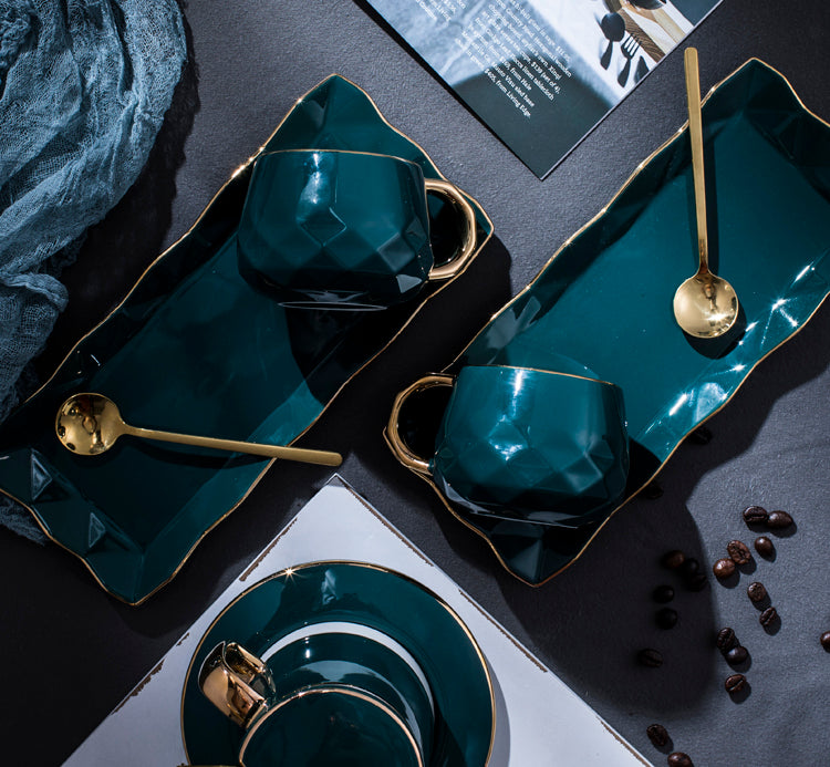 2 Sets of Emerald Green Dream Mug Set with Spoons on a table with gray table cloth, a magzine, a blanket and coffee beans strewn across the table. There is another mug and plate on the bottom right and it is a different emerald green and white mug set that is not available on the product listing.