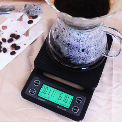 Backlit Electronic Scale with glass pour over coffee maker sitting on top of it. The coffee maker has water droplets due to condensation, in the inside of it and coffee grinders are visible on the top. In the background there are coffee beans sprawled across the counter.