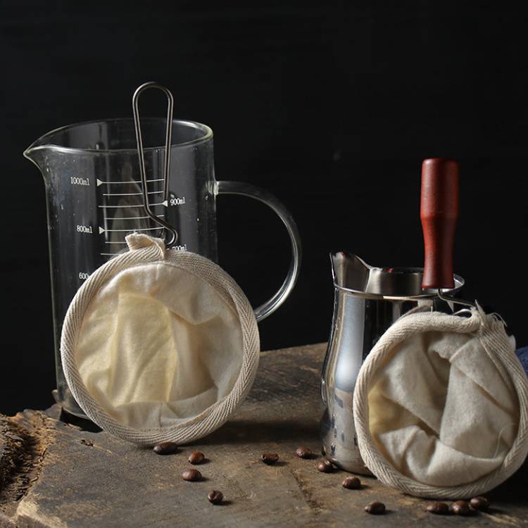 Reusable Cloth Coffee Filter with Stainless Steel Handle s and a milk frother jug, and a clear, glass measuring cup in the background. All on a table.