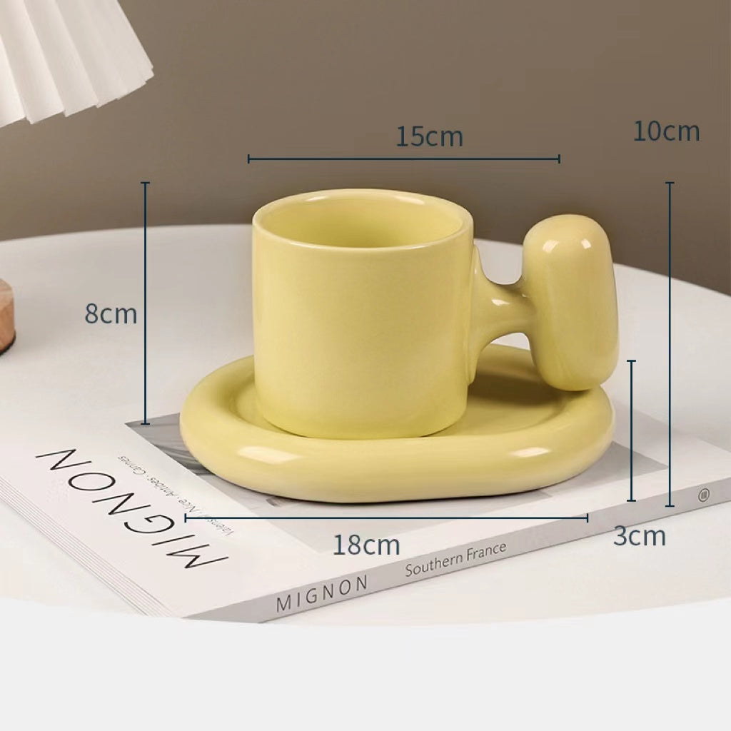 Fun and Bubbly Mug and Saucer Set in muted yellow on top of a book, on top on a table. There is a lamp shade on the top left of the photo. There are product dimensions shown on the photograph.